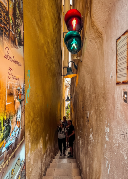 Very narrow, stepped alleyway between tall buildings with a traffic light for pedestrians (red and green figures), and tourists descending the stairs.