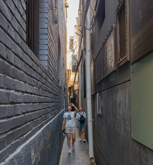 Two people walking down a dark, narrow street lined with tall, dark-grey brick walls and informational plaques in a Chinese city.