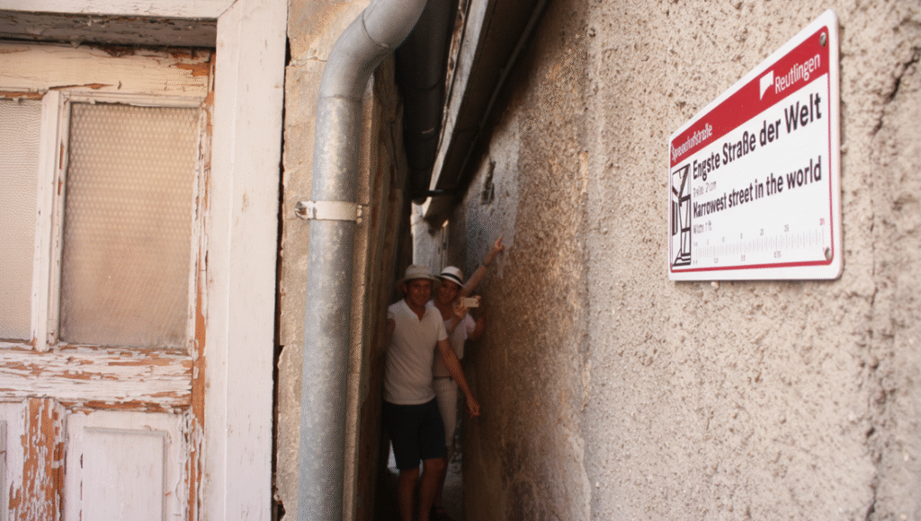 Two tourists standing in the world's narrowest street, the Spreuerhofstraße in Reutlingen, Germany, with a sign reading 'Engste Straße der Welt / Narrowest street in the world'.