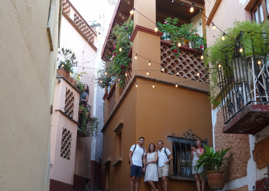 A small group of tourists posing for a photo in a charming, tight Callejón del Beso alley with balconies adorned with flowers and string lights.
